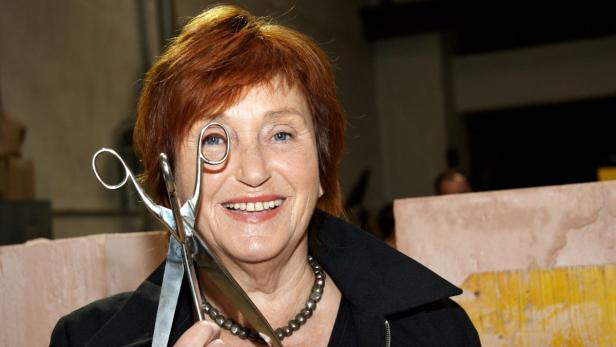 Eine Frau mit roten Haaren hält eine große Schere vor ihr Auge.