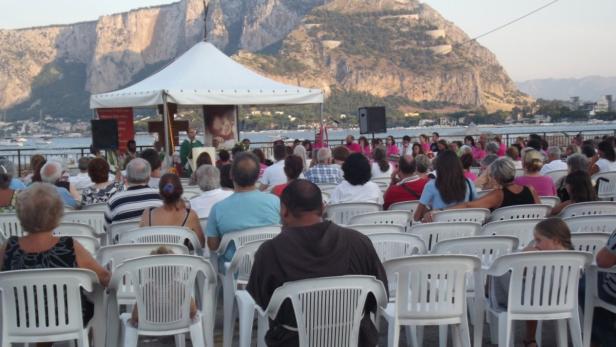 Eine Menschenmenge sitzt vor einer Bühne mit einem Priester und einem großen Berg im Hintergrund.