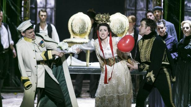 Auf einer Bühne streiten sich eine Frau mit Krone und rotem Ballon mit zwei Männern in Uniformen.