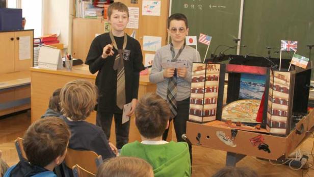 Schüler präsentieren ein Projekt mit einem Modellgebäude und Länderflaggen vor einer Klasse.