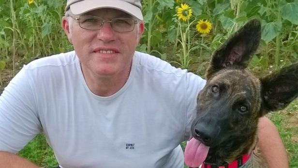 Ein Mann mit Brille und Kappe posiert mit seinem Hund vor einem Feld mit Sonnenblumen.