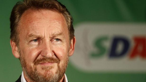 Nahaufnahme eines Mannes mit Bart vor einem grünen Hintergrund mit dem Schriftzug SDA.