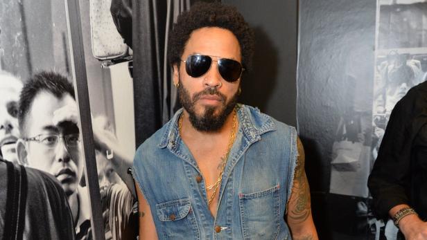 Lenny Kravitz mit Sonnenbrille und Jeansweste posiert vor einer Fotowand.
