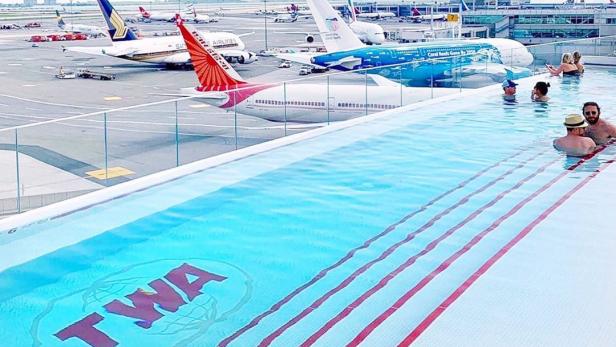 Menschen schwimmen in einem Pool mit Blick auf den Flughafen und Flugzeuge. Das TWA-Logo ist im Pool zu sehen.