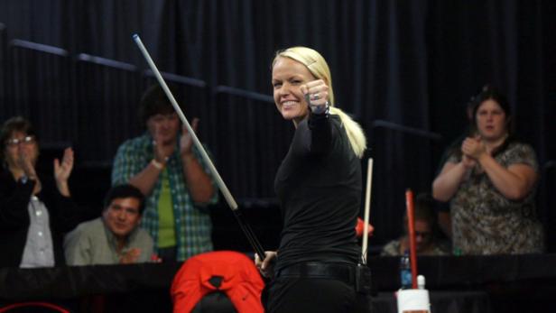 Eine blonde Billardspielerin jubelt mit Queue in der Hand vor applaudierenden Zuschauern.