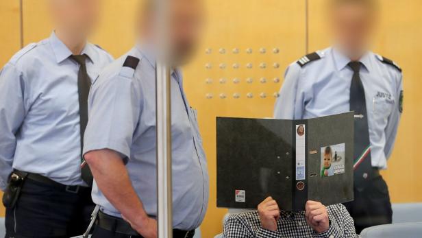 Ein Angeklagter versteckt sein Gesicht hinter einer Akte in einem Gerichtssaal, bewacht von Justizbeamten.