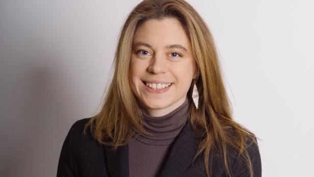 Eine lächelnde Frau mit blonden Haaren, einem braunen Rollkragenpullover und einem Blazer.