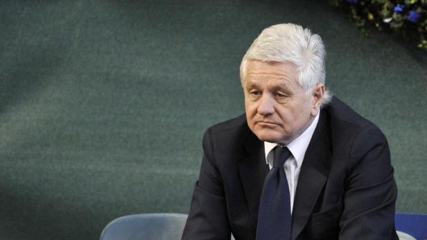 Ein Mann mit grauem Haar und dunklem Anzug sitzt vor einem grünen Hintergrund.