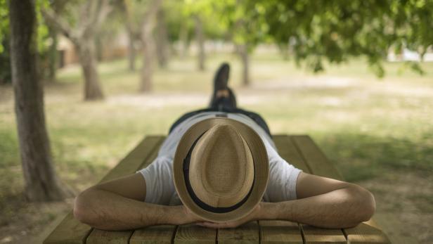 Eine Person liegt mit Hut entspannt auf einer Holzbank im grünen Park.