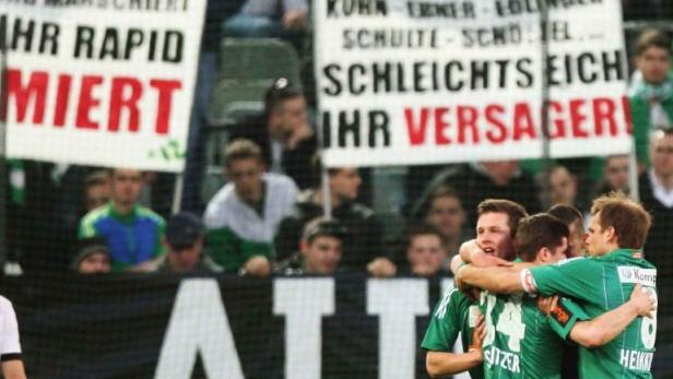 Jubelnde Fußballspieler vor Zuschauern mit Spruchbändern wie „Weil ihr Rapid blamiert“.