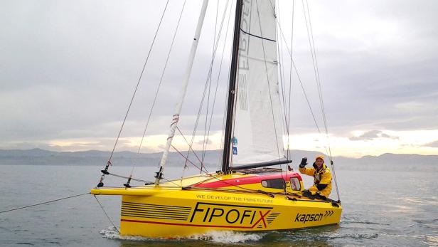Ein Mann in gelber Regenkleidung winkt von einem gelben Segelboot mit der Aufschrift „Fipofix“.