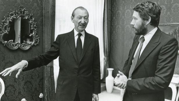 Zwei Männer in Anzügen unterhalten sich in einem eleganten Raum.