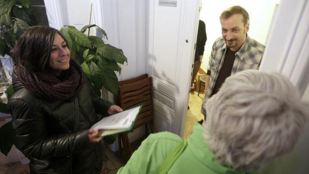Eine Frau mit Schal hält ein Dokument in der Hand, während ein Mann in der Tür steht.