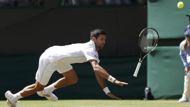 Novak Đoković rutscht auf dem Rasen, während ein Tennisball und sein Schläger in der Luft sind.