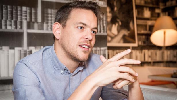 Ein Mann mit hellblauem Hemd gestikuliert vor einem Bücherregal.