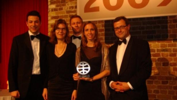 Eine Gruppe von Personen posiert mit einer Auszeichnung vor einer Backsteinmauer mit der Aufschrift „2009“.