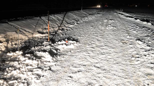 Eine verschneite Straße mit Reifenspuren und Markierungsstangen bei Nacht.