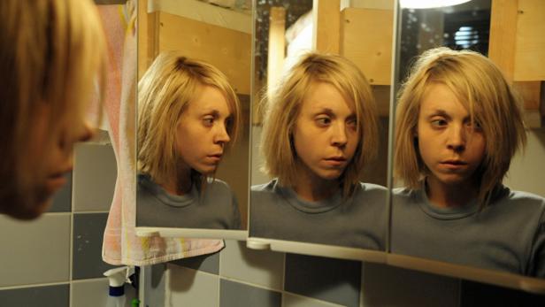 Eine junge Frau mit blonden Haaren betrachtet sich in einem dreiteiligen Spiegel im Badezimmer.