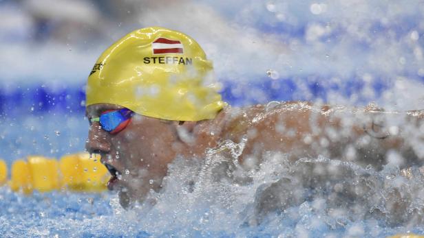 Ein Schwimmer mit gelber Badekappe und österreichischer Flagge im Gesicht schwimmt im Wasser.