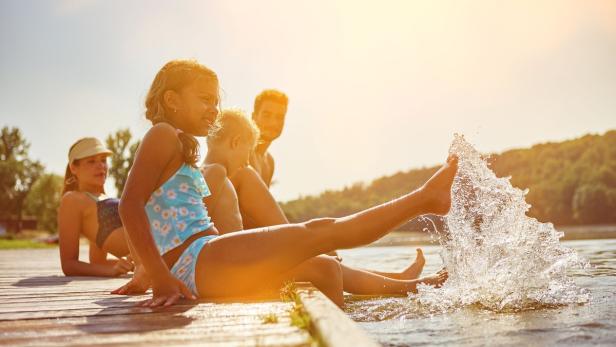 Eine Familie sitzt an einem sonnigen Tag auf einem Holzsteg am See und planscht mit den Füßen im Wasser.