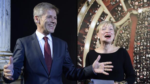 Ein Mann im Anzug und eine Frau stehen vor einem Hintergrund mit einem Opernhaus.