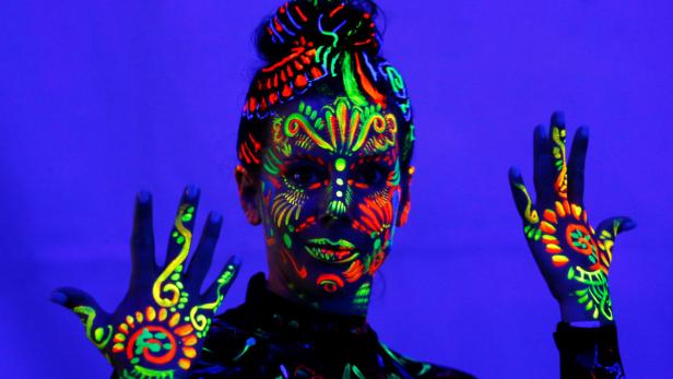 Eine Frau mit leuchtender Neonfarbe im Gesicht und auf den Händen vor blauem Hintergrund.