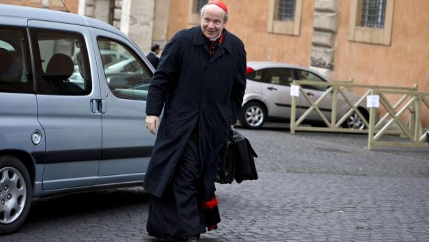 Ein Kardinal in schwarzer Kleidung und rotem Käppchen geht mit einer Aktentasche die Straße entlang.
