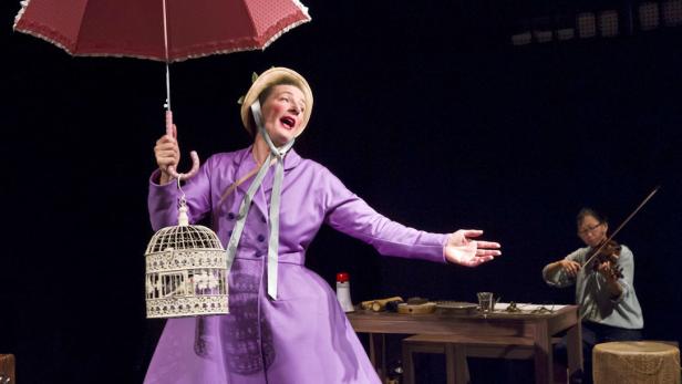 Eine Frau in einem lila Kostüm steht mit einem Vogelkäfig und einem Regenschirm auf einer Bühne, während eine Geigerin spielt.