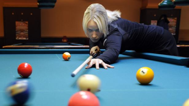 Eine blonde Frau zielt mit einem Billardqueue auf eine Billardkugel.