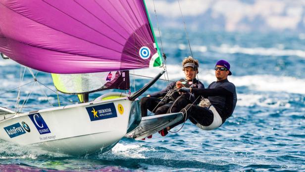 Zwei Segler steuern ein Segelboot mit lila Segel auf dem offenen Meer.