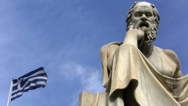 Eine Statue von Sokrates vor einer griechischen Flagge unter blauem Himmel.