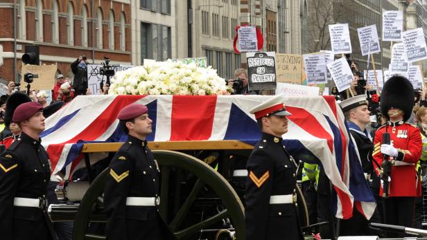 Ein Sarg, bedeckt mit der britischen Flagge und Blumen, wird von Soldaten getragen, während Demonstranten protestieren.