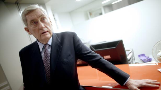 Ein älterer Mann im Anzug steht an einem orangefarbenen Schreibtisch.