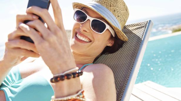 Eine Frau mit Hut und Sonnenbrille liegt am Pool und benutzt ihr Handy.