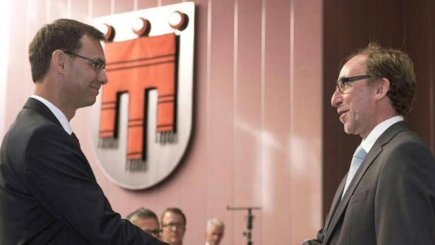 Zwei Männer in Anzügen schütteln sich vor einem Wappen die Hände.