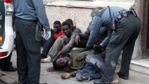 Einige erschöpfte afrikanische Migranten sitzen oder liegen auf dem Boden, während ein Polizist sie bewacht.