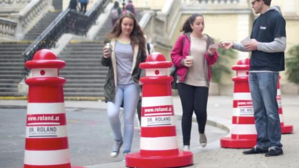 Zwei junge Frauen gehen an rot-weißen Verkehrspollern von Dr. Roland vorbei, während ein Mann Flyer verteilt.