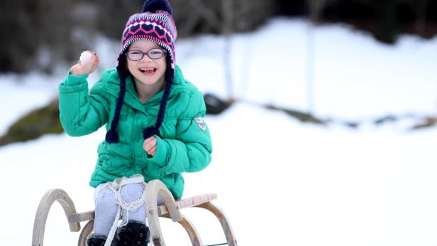 Ein Mädchen mit Brille und Mütze sitzt auf einem Schlitten und hält einen Schneeball in der Hand.