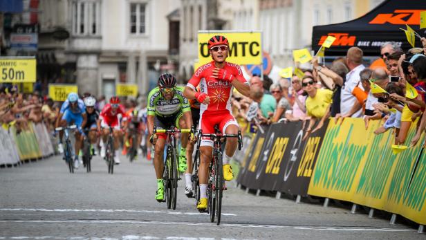 Ein Radrennfahrer überquert die Ziellinie, umjubelt von Zuschauern.