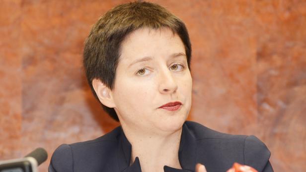 Eine Frau mit kurzem, dunklem Haar und dunklem Blazer vor einem orangefarbenen Hintergrund.