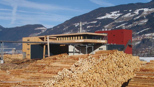 Ein Sägewerk mit großen Holzlagerflächen vor einer Bergkulisse.