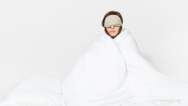 Fun tired young woman sitting in bed with sleep mask, sheet, pillow, wrapping in blanket isolated on white background. Beauty female spending time in room. Rest, relax, good mood concept. Copy space.