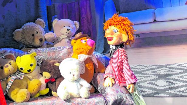 Eine Gruppe von Teddybären und eine Puppe sitzen auf einer Couch vor einem gemalten Hintergrund.