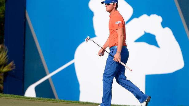 Ein Golfer in orangefarbenem Hemd und blauer Hose geht mit einem Putter über das Grün.
