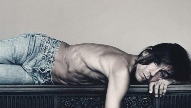Iggy Pop liegt mit nacktem Oberkörper und Jeans auf einem verzierten Möbelstück.