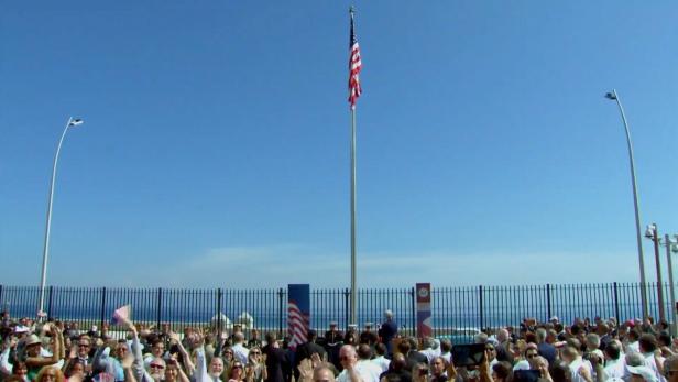 Eine Menschenmenge versammelt sich unter einer US-Flagge, die an einem sonnigen Tag weht.