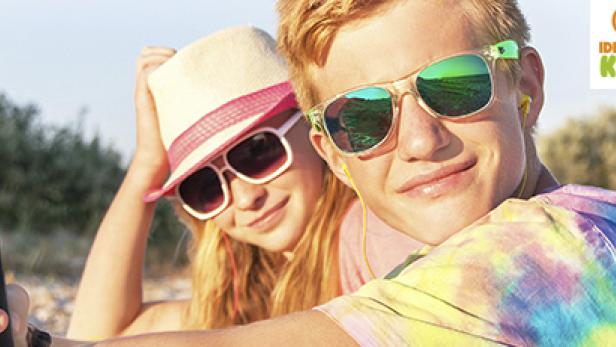 Zwei Jugendliche mit Sonnenbrillen und Kopfhörern hören Musik am Strand.
