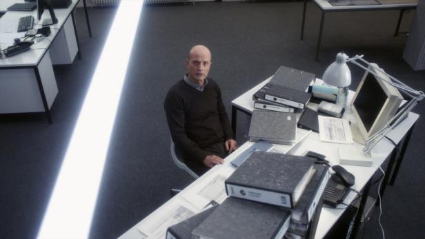 Ein Mann sitzt an einem Schreibtisch voller Aktenordner in einem Büro.