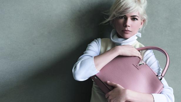 Michelle Williams hält eine rosafarbene Handtasche vor einem grauen Hintergrund.