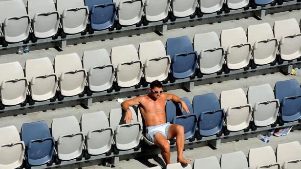 Ein Mann sitzt mit freiem Oberkörper und Sonnenbrille auf der Tribüne eines Stadions.